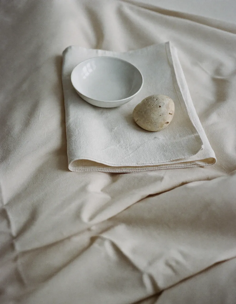 A folded ivory towel, a plain ceramic bowl and a smooth pale stone on pale oyster linen